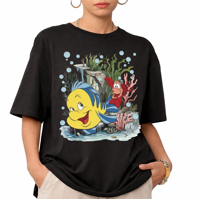Puede incluir: Camiseta negra con un gr&aacute;fico colorido de Flounder, Sebasti&aacute;n y un paisaje submarino de La Sirenita. El dise&ntilde;o incluye burbujas y coral. La camiseta tiene un ajuste casual y holgado.