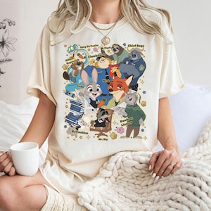 Op de afbeelding: Cr&egrave;mekleurig T-shirt met een kleurrijke cartoonafbeelding van personages uit de film Zootopia. De afbeelding bevat Judy Hopps, Nick Wilde en andere personages met hun namen. Het shirt heeft een ronde hals en korte mouwen.