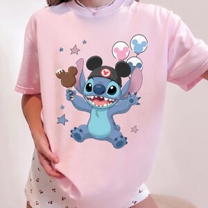May include: A light pink t-shirt featuring a graphic of the blue character Stitch wearing black mouse ears with a red emblem. Stitch holds a brown mouse-shaped ice cream bar and is surrounded by three circular balloons and several small stars.