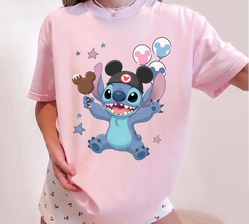 May include: A light pink t-shirt featuring a graphic of the blue character Stitch wearing black mouse ears with a red emblem. Stitch holds a brown mouse-shaped ice cream bar and is surrounded by three circular balloons and several small stars.