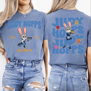 May include: A blue t-shirt featuring a graphic of the character Judy Hopps from Zootopia. The front displays the text Judy Hopps, Est. 2016, and Zootopia. The back shows a larger Judy Hopps graphic with repeating text, a donut, a carrot, and a badge.