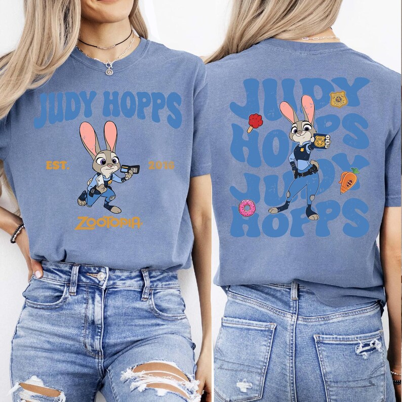 May include: A blue t-shirt featuring a graphic of the character Judy Hopps from Zootopia. The front displays the text Judy Hopps, Est. 2016, and Zootopia. The back shows a larger Judy Hopps graphic with repeating text, a donut, a carrot, and a badge.