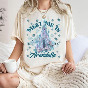 May include: Cream-colored t-shirt featuring a graphic of a castle with the text "Meet Me In Arendelle." The castle design incorporates shades of blue, purple, and pink, with surrounding snowflakes and mountains. The shirt is short-sleeved.