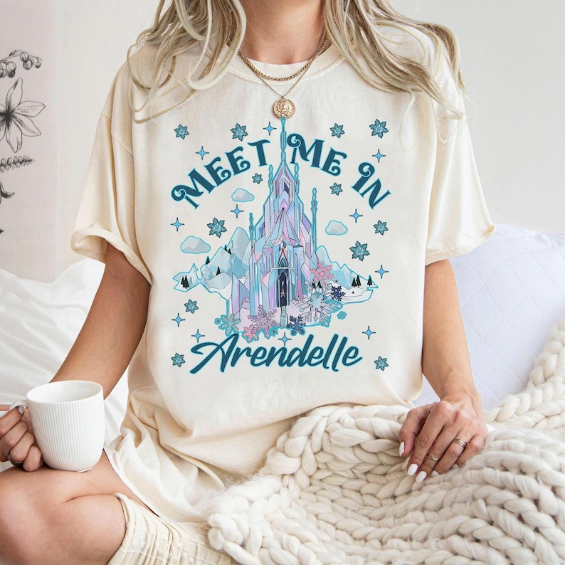 May include: Cream-colored t-shirt featuring a graphic of a castle with the text "Meet Me In Arendelle." The castle design incorporates shades of blue, purple, and pink, with surrounding snowflakes and mountains. The shirt is short-sleeved.