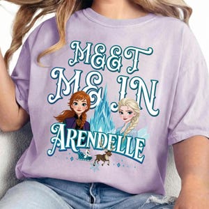 May include: A light purple t-shirt featuring the text "MEET ME IN ARENDELLE" in teal script. The graphic includes illustrations of Anna and Elsa from Frozen, along with a reindeer and a sleigh.