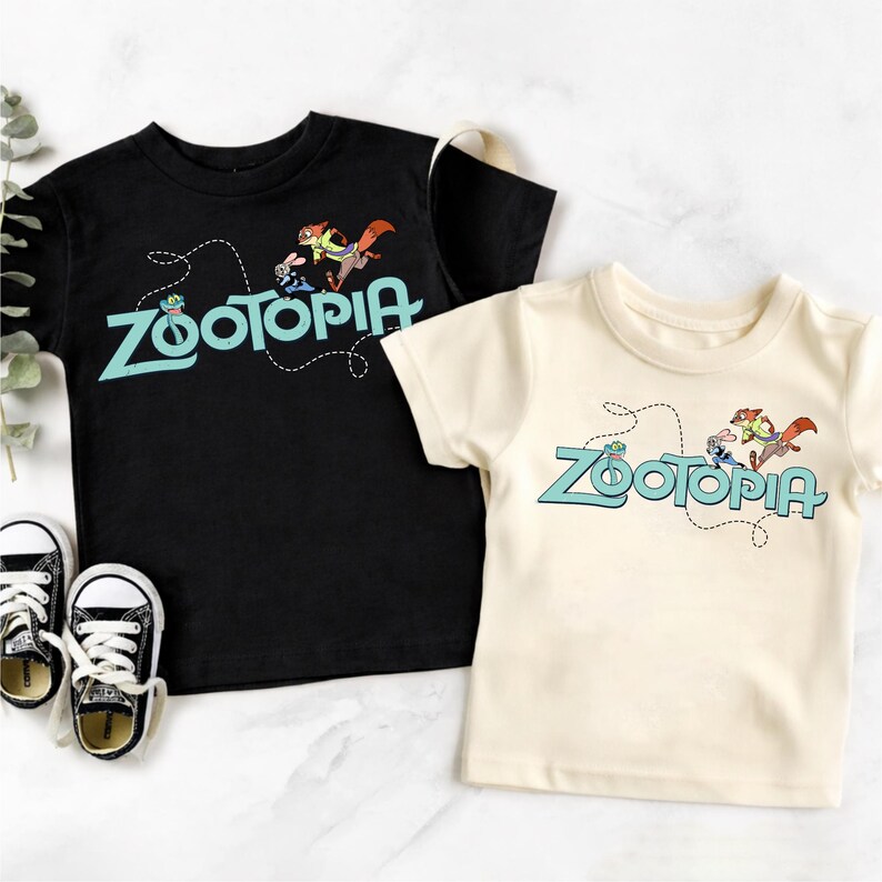 May include: A black t-shirt and a cream t-shirt, each featuring the Zootopia logo in light blue with a dashed line path and cartoon characters. The black shirt is paired with a black and white sneaker, while the cream shirt is plain.