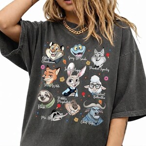 May include: A charcoal gray short-sleeve t-shirt featuring a graphic print of various animated animal characters including Judy Hopps, Nick Wilde, Flash, and Chief Bogo, with their names printed in white script text alongside small colorful icons.
