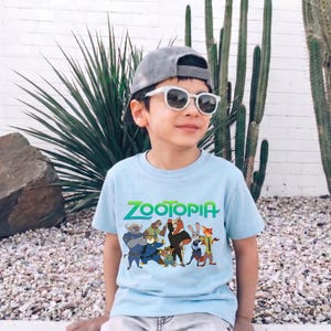 K&ouml;nnte beinhalten: Hellblaues T-Shirt mit dem Wort "ZOOTOPIA" in Gr&uuml;n und einer Grafik von Charakteren aus dem Animationsfilm. Das T-Shirt wird von einer Person getragen, die eine graue Kappe und eine Sonnenbrille tr&auml;gt.