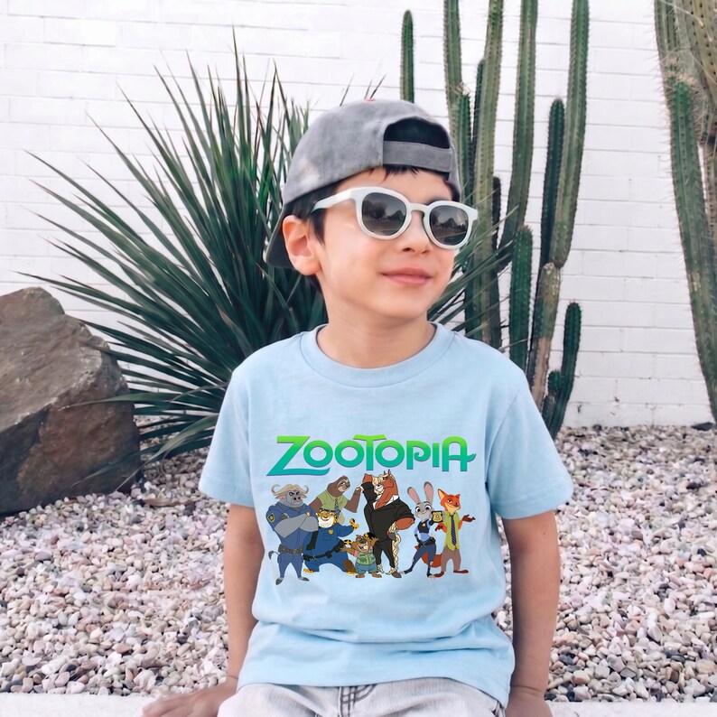 K&ouml;nnte beinhalten: Hellblaues T-Shirt mit dem Wort "ZOOTOPIA" in Gr&uuml;n und einer Grafik von Charakteren aus dem Animationsfilm. Das T-Shirt wird von einer Person getragen, die eine graue Kappe und eine Sonnenbrille tr&auml;gt.