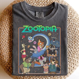 May include: A charcoal gray crew neck t-shirt featuring a colorful graphic print of Zootopia characters including a fox, rabbit, sloth, and snake. The design includes the text Zootopia in a bright green font, surrounded by small stars and icons.