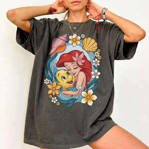 May include: Dark gray t-shirt featuring a colorful illustration of Ariel and Flounder from The Little Mermaid. The design is framed by flowers and seashells. The shirt has a relaxed fit and short sleeves.
