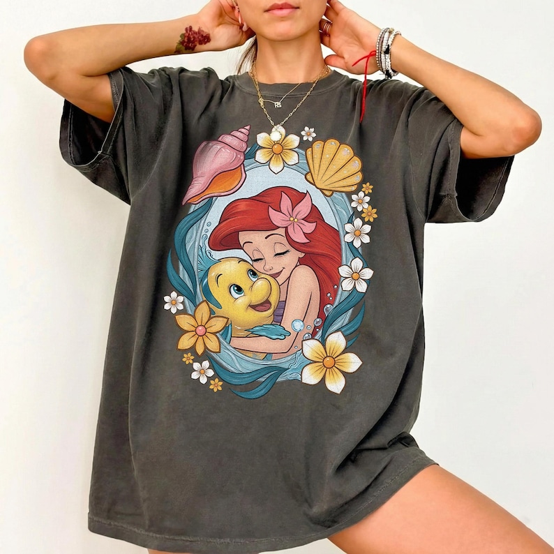 May include: Dark gray t-shirt featuring a colorful illustration of Ariel and Flounder from The Little Mermaid. The design is framed by flowers and seashells. The shirt has a relaxed fit and short sleeves.
