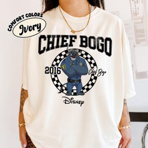 May include: An ivory t-shirt featuring a graphic of Chief Bogo from Disney's Zootopia. The design includes the text CHIEF BOGO, 2016, and Disney, with a black and white checkered circle surrounding the character. A Comfort Colors Ivory label is visible.