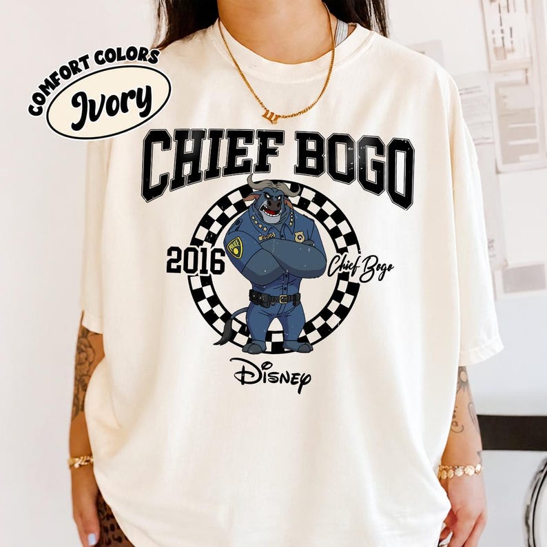 May include: An ivory t-shirt featuring a graphic of Chief Bogo from Disney's Zootopia. The design includes the text CHIEF BOGO, 2016, and Disney, with a black and white checkered circle surrounding the character. A Comfort Colors Ivory label is visible.