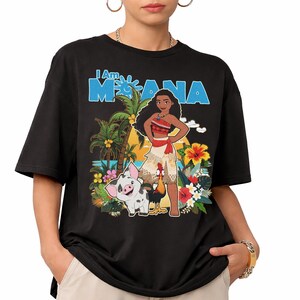 May include: Black t-shirt featuring a colorful graphic of Moana, Pua the pig, and Hei Hei the rooster. The text "I Am Moana" is printed in blue. The design includes tropical flowers, palm trees, and a beach scene.