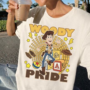 May include: Cream-colored t-shirt featuring a graphic of Woody from Toy Story. The design includes the word "WOODY" in yellow block letters, Woody's image, and the word "PRIDE" in brown block letters. Additional graphics include stars, a ball, and a hat.