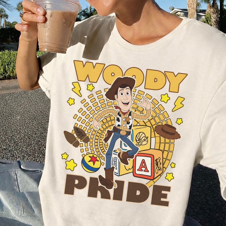 May include: Cream-colored t-shirt featuring a graphic of Woody from Toy Story. The design includes the word "WOODY" in yellow block letters, Woody's image, and the word "PRIDE" in brown block letters. Additional graphics include stars, a ball, and a hat.