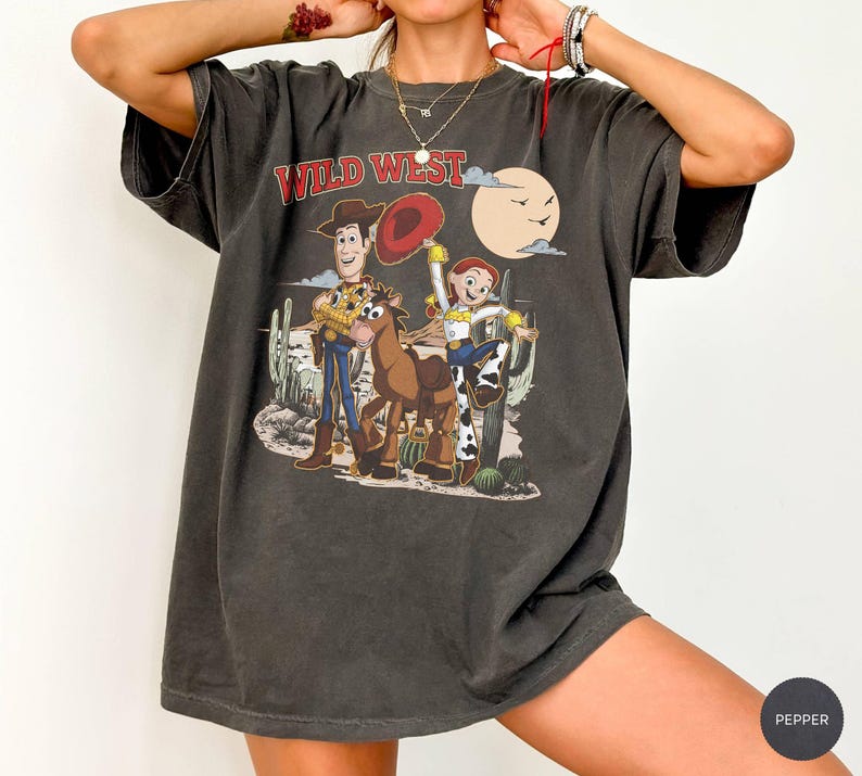 Pu&ograve; includere: T-shirt grigio scuro con un design del Far West con i personaggi dei cartoni animati Woody, Jessie e un cavallo. La grafica include una scena desertica con cactus e una luna piena. La maglietta ha un aspetto vintage.