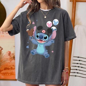 May include: A charcoal gray short-sleeve t-shirt featuring a graphic of the blue character Stitch wearing a black hat with ears. Stitch holds a brown ice cream bar and is surrounded by three round balloons and small pink and blue stars.
