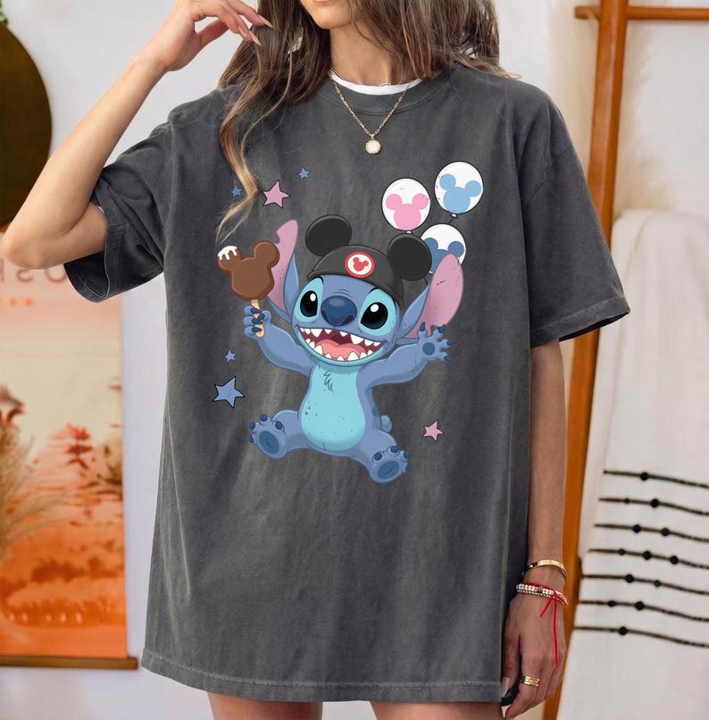 May include: A charcoal gray short-sleeve t-shirt featuring a graphic of the blue character Stitch wearing a black hat with ears. Stitch holds a brown ice cream bar and is surrounded by three round balloons and small pink and blue stars.
