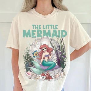 May include: Cream-colored t-shirt featuring a graphic of Ariel from The Little Mermaid. The design includes Ariel in a seashell, with Flounder and Sebastian. The text "THE LITTLE MERMAID" is above the image in teal.