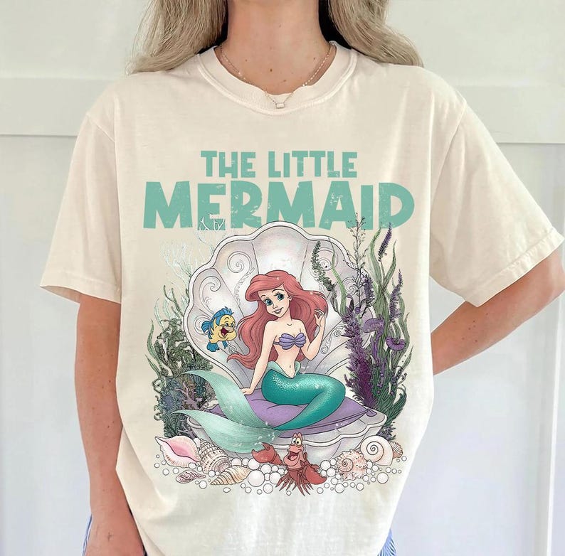 May include: Cream-colored t-shirt featuring a graphic of Ariel from The Little Mermaid. The design includes Ariel in a seashell, with Flounder and Sebastian. The text "THE LITTLE MERMAID" is above the image in teal.