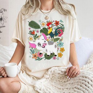 Puede incluir: Camiseta color crema con un dise&ntilde;o de cerdo y gallo de dibujos animados, rodeados de flores y hojas coloridas. El cerdo tiene orejas rosas y una cara sonriente. El gallo tiene plumas rojas, amarillas y azules. El dise&ntilde;o est&aacute; sobre un fondo floral.
