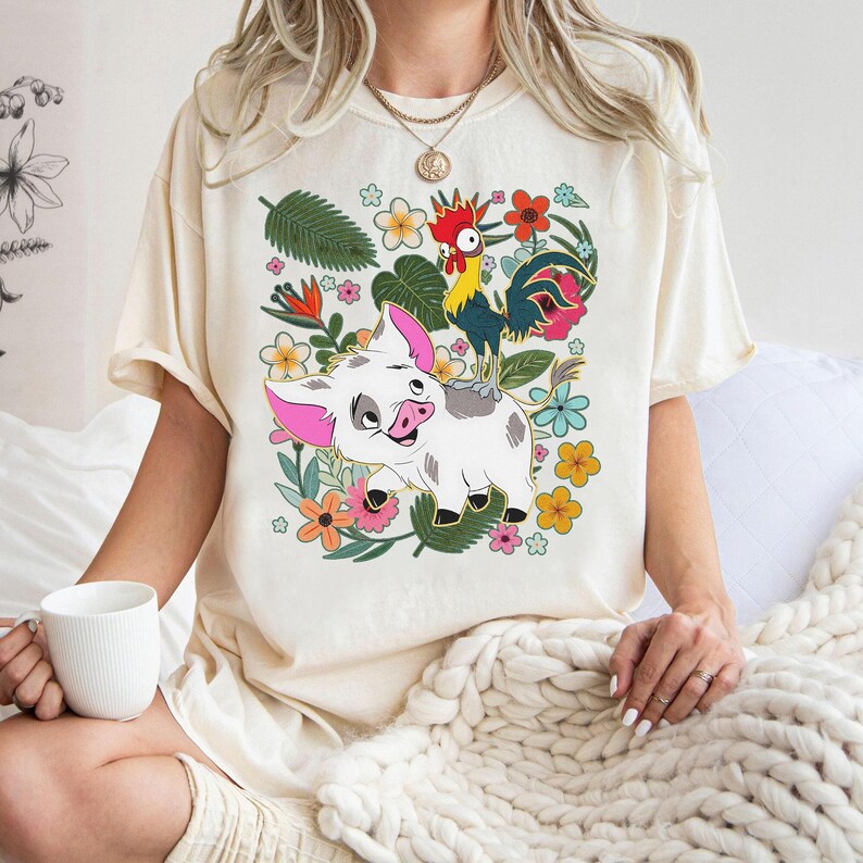 Puede incluir: Camiseta color crema con un dise&ntilde;o de cerdo y gallo de dibujos animados, rodeados de flores y hojas coloridas. El cerdo tiene orejas rosas y una cara sonriente. El gallo tiene plumas rojas, amarillas y azules. El dise&ntilde;o est&aacute; sobre un fondo floral.