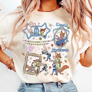 May include: An ivory t-shirt featuring a graphic print of Zootopia characters Judy Hopps and Nick Wilde. The design includes multiple illustrations of the characters, floral accents, bows, and the text Welcome to Zootopia and Zootopia.