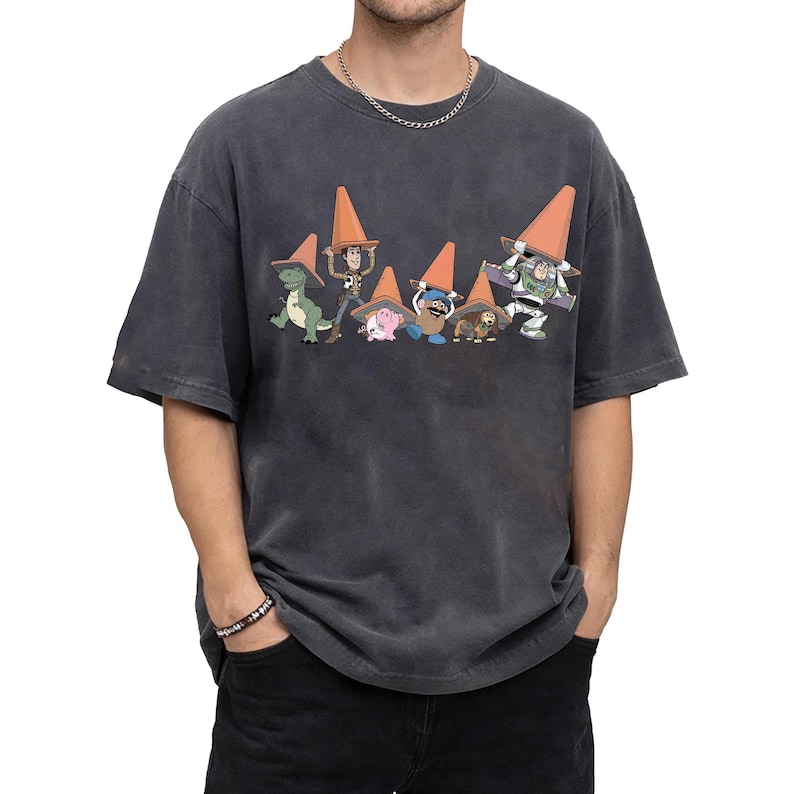 May include: Dark gray t-shirt featuring a graphic of Toy Story characters wearing orange traffic cones. The characters include Rex, Woody, Hamm, Mr. Potato Head, Slinky Dog, and Buzz Lightyear. The shirt has a vintage, washed look.