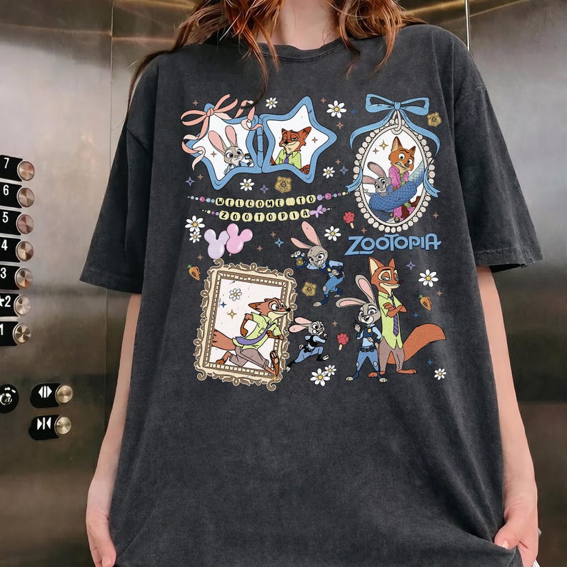 May include: A charcoal gray short-sleeve t-shirt featuring a graphic print of characters from Zootopia. The design includes framed illustrations of Judy Hopps and Nick Wilde, floral accents, and the text Welcome to Zootopia and Zootopia.