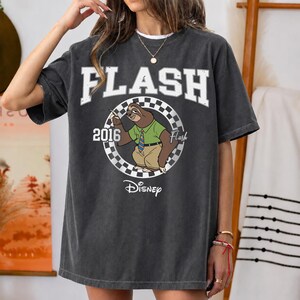 May include: A charcoal grey short-sleeve t-shirt featuring a graphic of the character Flash the sloth from Disney. The design includes the text FLASH, 2016, and Disney, with a checkered circular border surrounding the sloth wearing a shirt and tie.