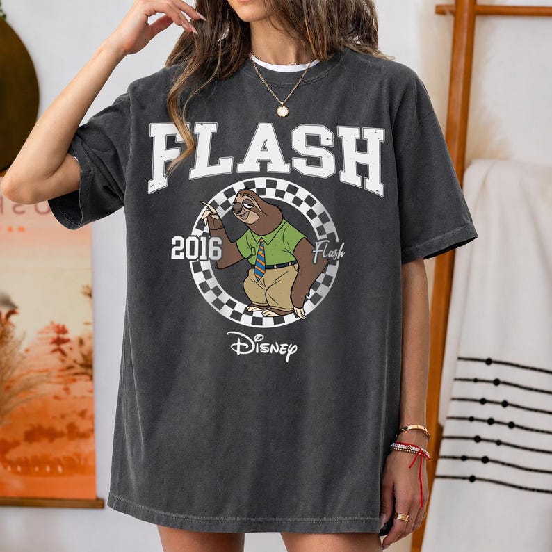 May include: A charcoal grey short-sleeve t-shirt featuring a graphic of the character Flash the sloth from Disney. The design includes the text FLASH, 2016, and Disney, with a checkered circular border surrounding the sloth wearing a shirt and tie.