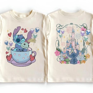 May include: A cream-colored t-shirt shown from the front and back. The front features a blue alien character sitting in a light purple teacup with hearts. The back displays the same character facing a large castle with fireworks and floral accents.