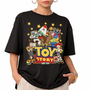 May include: Black t-shirt featuring a colorful graphic of Toy Story characters, including Woody, Buzz Lightyear, and others. The words "TOY STORY" are in yellow, with "SINCE 1995" below. The shirt has short sleeves and a crew neck.