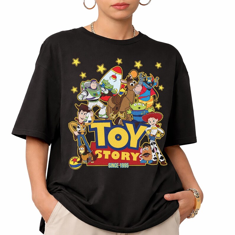 May include: Black t-shirt featuring a colorful graphic of Toy Story characters, including Woody, Buzz Lightyear, and others. The words "TOY STORY" are in yellow, with "SINCE 1995" below. The shirt has short sleeves and a crew neck.