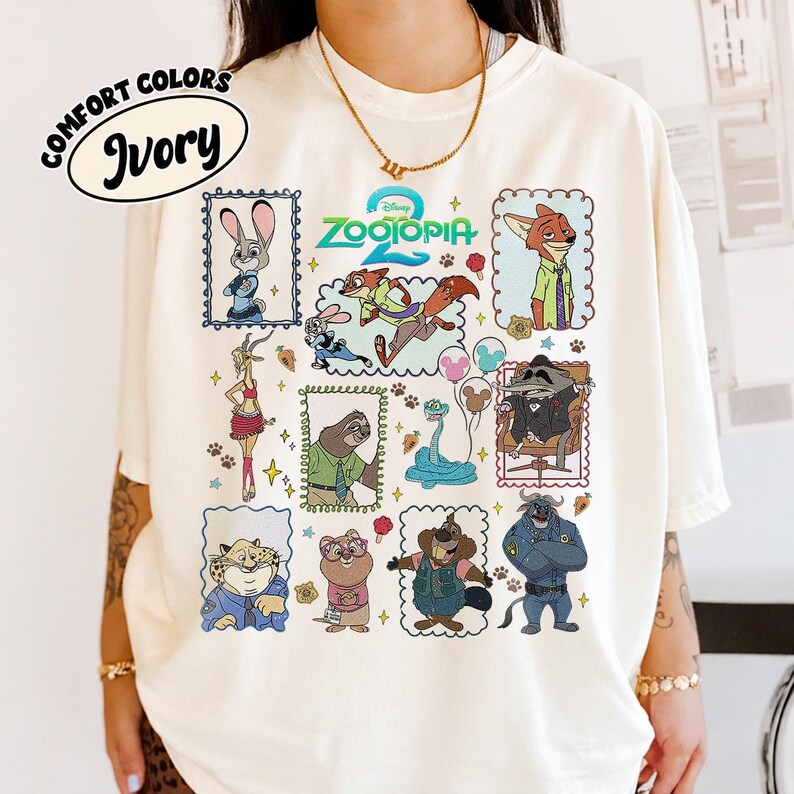 May include: An ivory t-shirt featuring a colorful graphic print of various Zootopia characters including Judy Hopps, Nick Wilde, Flash, and Chief Bogo arranged in individual frames, with the Zootopia 2 logo centered at the top of the design.