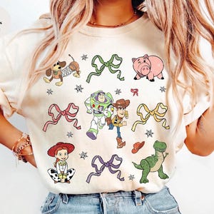 Op de afbeelding: Ivoor kleurig T-shirt met kleurrijke cartoonfiguren en strikken. Het ontwerp bevat een hond, een varken, Buzz Lightyear, Woody, Jessie en een dinosaurus, samen met sterren en strikken. De tekst "Colors Ivory" staat linksboven.