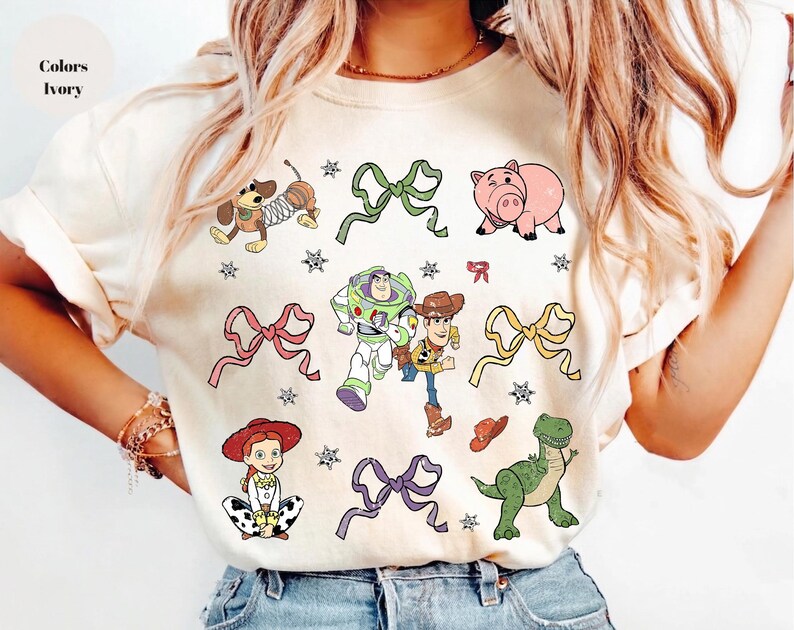 Op de afbeelding: Ivoor kleurig T-shirt met kleurrijke cartoonfiguren en strikken. Het ontwerp bevat een hond, een varken, Buzz Lightyear, Woody, Jessie en een dinosaurus, samen met sterren en strikken. De tekst "Colors Ivory" staat linksboven.