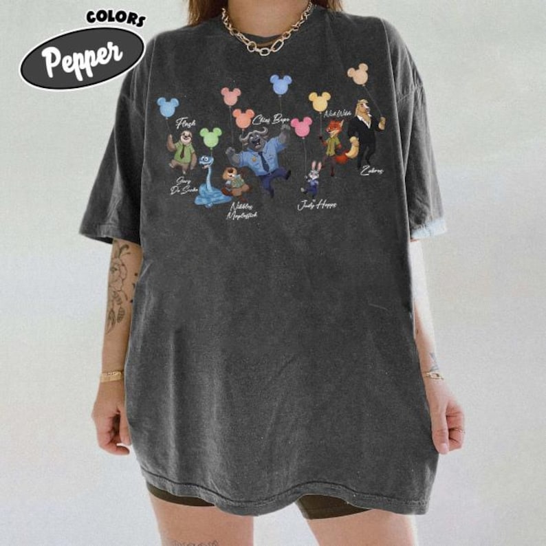 May include: A charcoal grey short-sleeve t-shirt featuring a graphic print of various animated animal characters, each holding a colourful balloon. The text includes names such as Flash, Chief Bogo, Nick Wilde, and Judy Hopps printed near the characters.