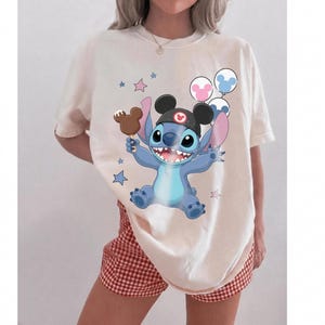 May include: A cream-colored t-shirt featuring a graphic of the blue character Stitch wearing black mouse ears, holding a brown chocolate-covered ice cream bar, and surrounded by small blue and pink stars and three round balloons.