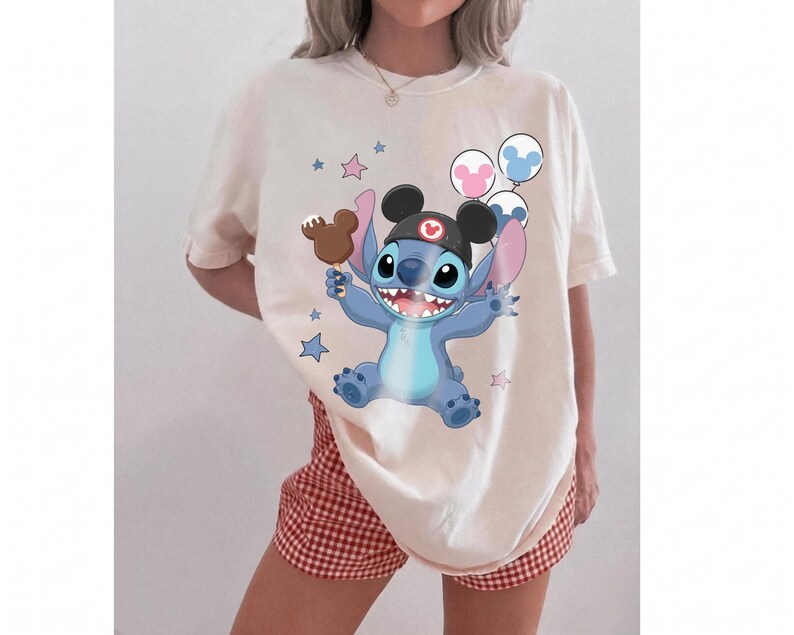 May include: A cream-colored t-shirt featuring a graphic of the blue character Stitch wearing black mouse ears, holding a brown chocolate-covered ice cream bar, and surrounded by small blue and pink stars and three round balloons.