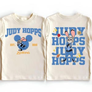 May include: A cream-colored t-shirt featuring Judy Hopps from Zootopia. The front displays the text Judy Hopps, Est. 2016, and Zootopia with a blue Mickey Mouse silhouette. The back shows Judy Hopps with the text Judy Hopps, a carrot, and a badge.