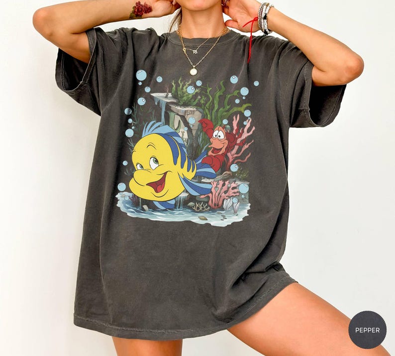Puede incluir: Camiseta gris oscuro con una ilustraci&oacute;n de dibujos animados de Flounder y Sebasti&aacute;n de La Sirenita. El dise&ntilde;o incluye burbujas azules, coral y algas marinas. La camiseta tiene mangas cortas y un corte holgado.