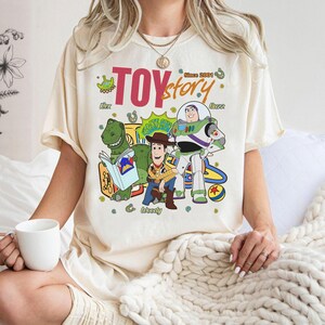 May include: Cream-colored t-shirt featuring a colorful graphic of Toy Story characters, including Rex, Woody, and Buzz Lightyear. The text "TOY story" is displayed above the characters, with "Since 2001" printed above. The shirt has short sleeves.
