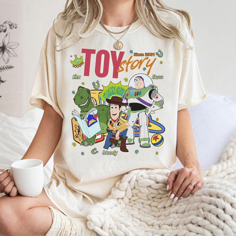 May include: Cream-colored t-shirt featuring a colorful graphic of Toy Story characters, including Rex, Woody, and Buzz Lightyear. The text "TOY story" is displayed above the characters, with "Since 2001" printed above. The shirt has short sleeves.
