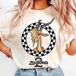 May include: An ivory t-shirt featuring a graphic of a gazelle with long horns and blonde hair inside a black and white checkered circle. Below the circle, the text Zootopia 2 is printed in black. The shirt has short sleeves and a crew neckline.
