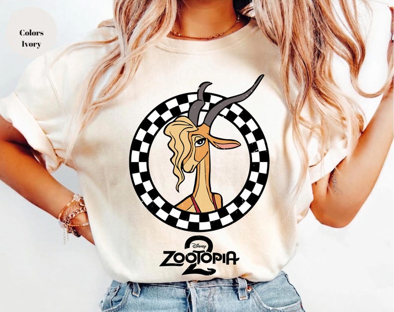 May include: An ivory t-shirt featuring a graphic of a gazelle with long horns and blonde hair inside a black and white checkered circle. Below the circle, the text Zootopia 2 is printed in black. The shirt has short sleeves and a crew neckline.