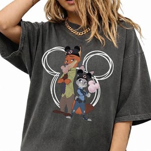 May include: A charcoal gray short-sleeve t-shirt featuring a graphic print of Nick Wilde and Judy Hopps from Zootopia. The characters are wearing black mouse ear hats and holding pink cotton candy, centered within a white circular outline.
