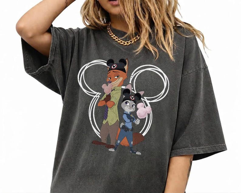 May include: A charcoal gray short-sleeve t-shirt featuring a graphic print of Nick Wilde and Judy Hopps from Zootopia. The characters are wearing black mouse ear hats and holding pink cotton candy, centered within a white circular outline.